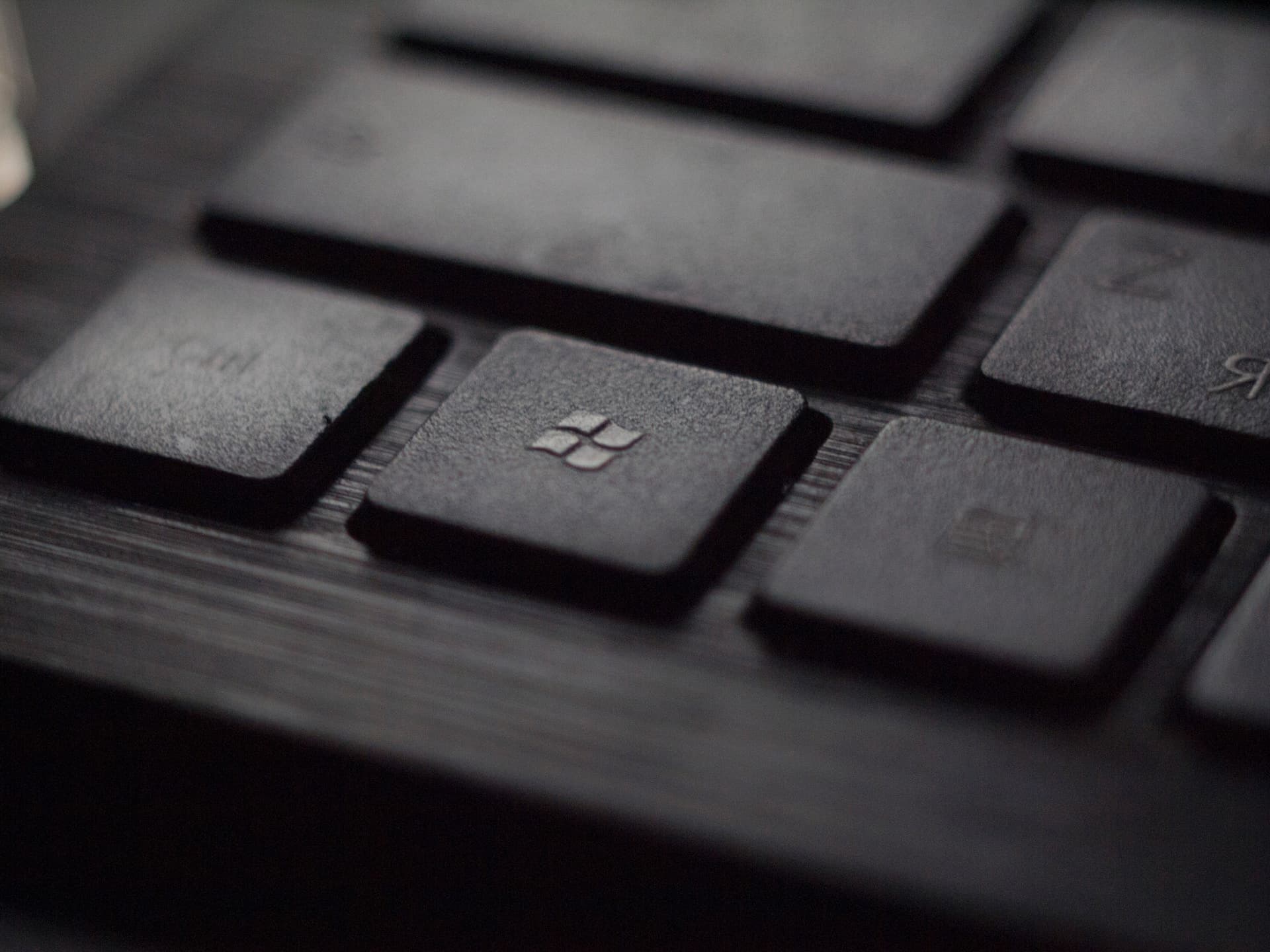 Close-up photograph of a black computer keyboard with shallow depth of field focusing on the Windows key in the center, which displays the older four-pane Windows logo. The surrounding keys are blurred, with the Ctrl key partially visible on the left and the Alt key area on the right. The image has a dark, moody aesthetic with muted gray tones.