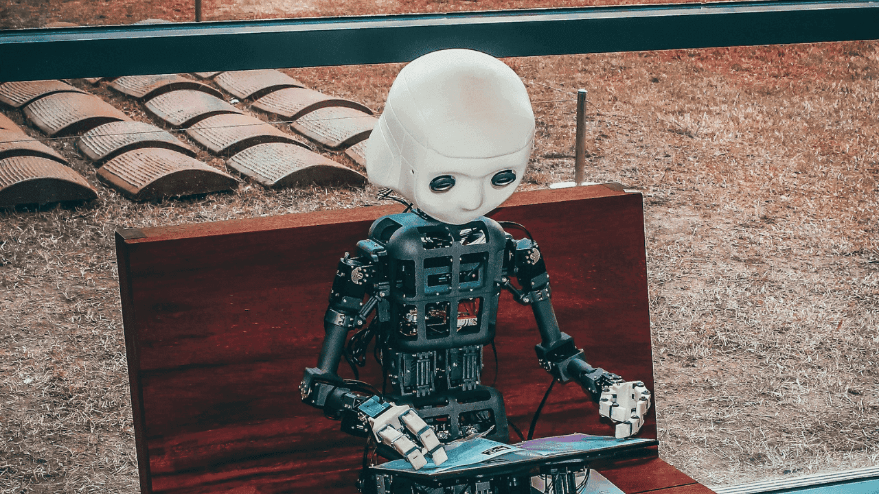 Blog thumbnail showing a small humanoid robot with a white head and exposed mechanical body seated on a wooden bench, looking down at and interacting with a tablet device placed on its lap, with an outdoor setting in the background featuring gravel ground and curved architectural elements.